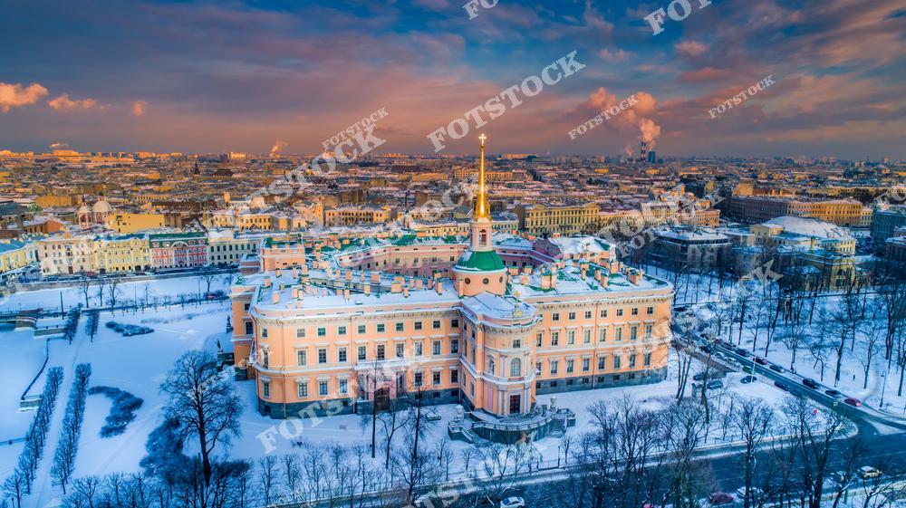 На фотографии изображён Михайловский (Инженерный) замок — уникальное архитектурное сооружение XVIII века, построенное по заказу Павла I. Комплекс расположен в центре Санкт-Петербурга, окружённый каналами и парками. Снег покрывает крыши и аллеи, придавая пейзажу особую торжественность. Вдали видна застройка исторического центра, освещённая мягкими красками закатного неба. Снимок сделан с воздуха и показывает гармонию архитектуры и природы.